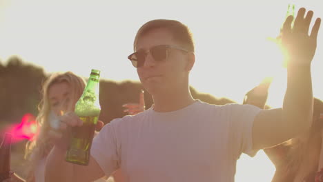 Young-man-in-white-T-shirt-and-black-fashionable-sun-glasses-is-dancing-with-his-best-friends-on-the-open-air-party-with-beer.-He-enjoys-this-summer-evening-at-sunset.